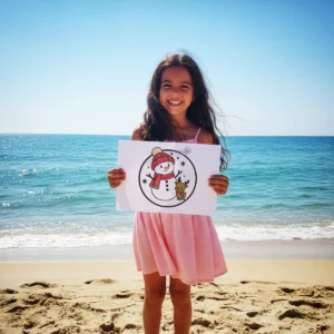 A smiling child standing on a sunny beach next to the ocean, proudly holding up a completed coloring page featuring a snowman and a reindeer.